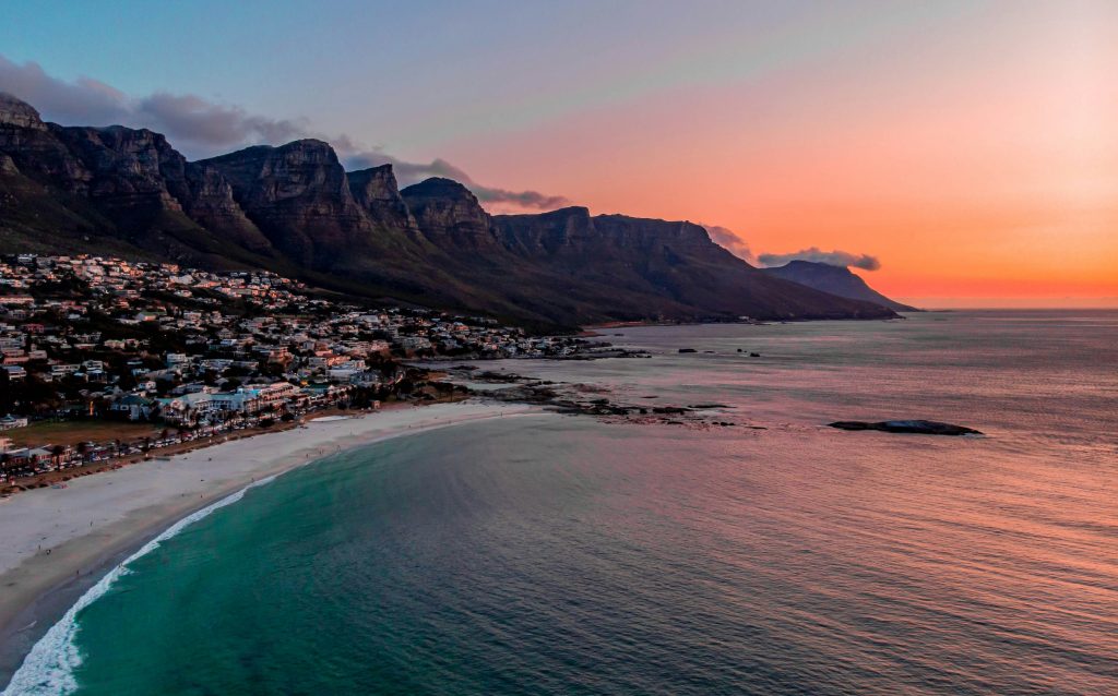 Vue panoramique sur Camps Bay à Cape Town, entre plage de sable blanc, palmiers et montagnes en arrière-plan, au coucher du soleil.